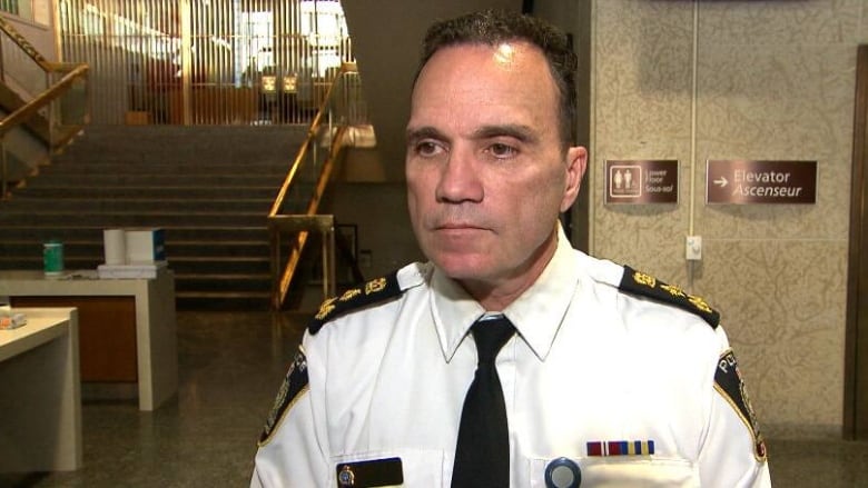 A man with short dark hair, wearing a white shirt with patches on the shoulders, stands in the lobby of a building.