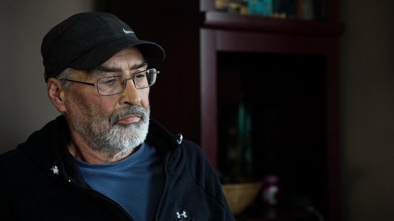 A man sits on a chair wearing a blue t-shirt, black sweater and black ball cap. 