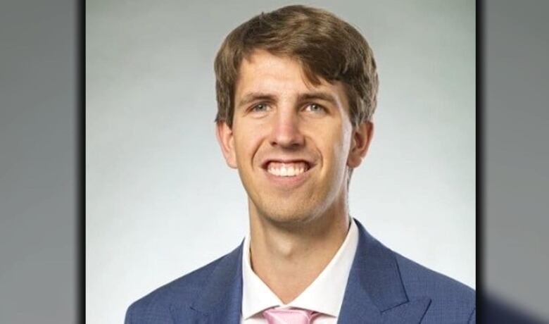 A man with short brown hair in a blue blazer and pink tie smiles for the camera.