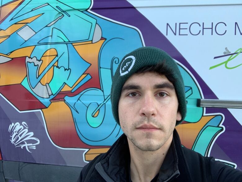 A young man with a toque stands in front of a wall mural