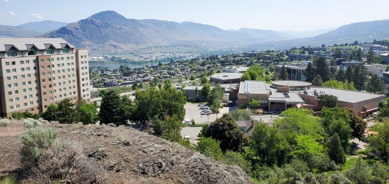 A panoramic view of a riverfront city with mountains in the background, with a university campus in the foreground.