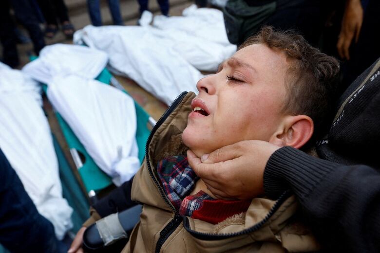 A crying child is held at a funeral as white body bags are seen in the background. 