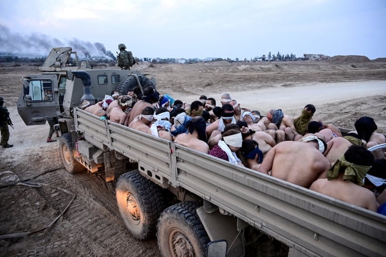 A large group of men are seen bound, topless and blindfolded in the back of a truck.