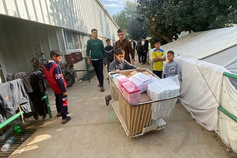 A child pushes a trolley with boxes in it, while other children and adults follow. 