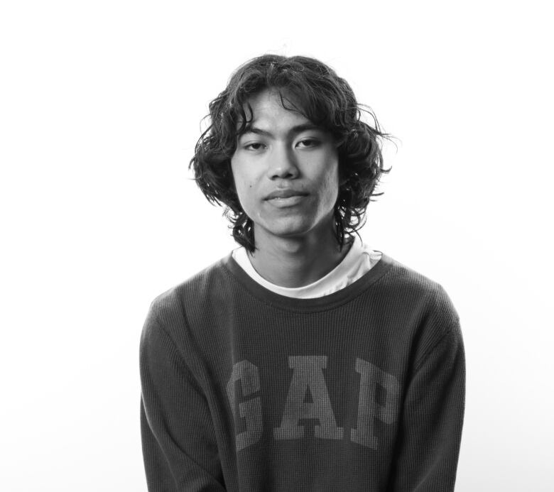 Black and white portrait of young man sitting on stool. He has dark, shaggy hair and a non-expressive look on his face. He is wearing a sweatshirt with Gap written across the front. 