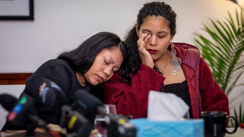 Two Indigenous women console each other as they sit in front of microphones at a press conference.