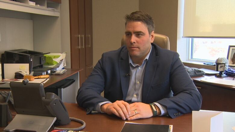 A man wearing a blue blazer over a light blue shirt is picture while sitting at a desk during an interview with a reporter.