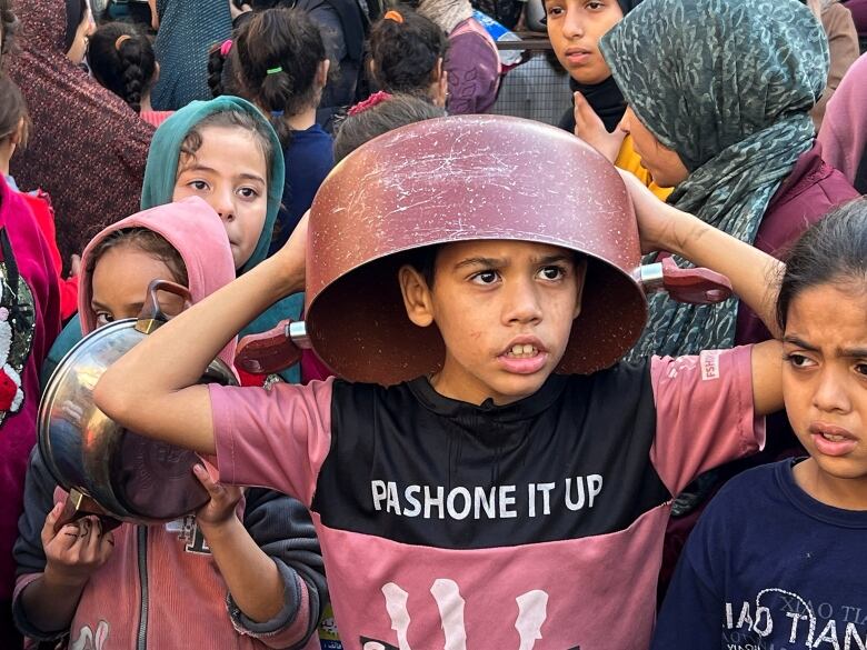 A young boy stands holding an empty pot over his head. He's among a crowd of other children, who are also holding pots.