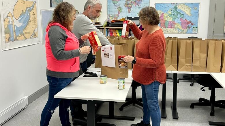 Three people seen standing in front of a line of paper bags while they pack food into them.