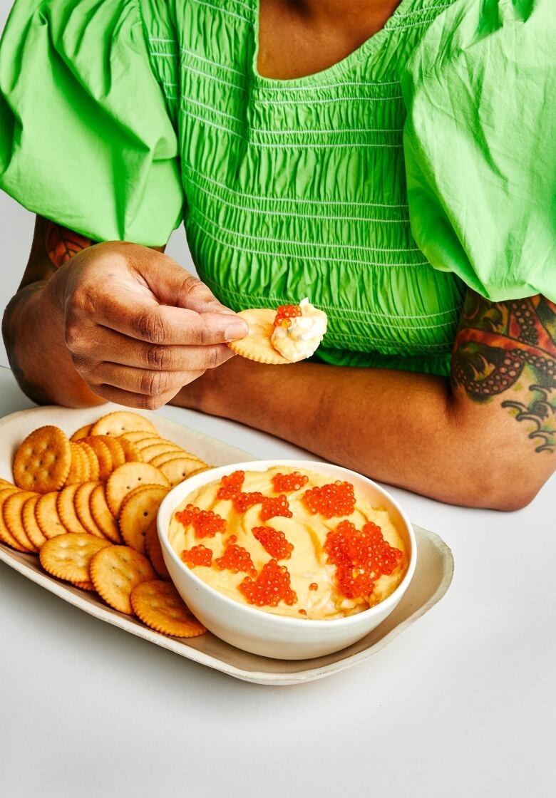 A bowl of light yellow dip with orange trout roe on top. The bowl is sitting on a platter with Ritz crackers next to it. Behind it, a person wearing a lime green dress scoops into  the dip with a cracker.  