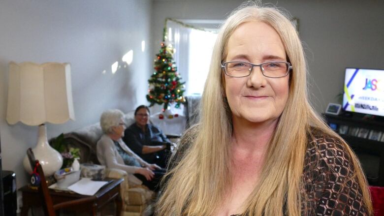 A woman poses for a photo in a private apartment building with her elderly motherly sitting with her home care worker on a couch in the background.