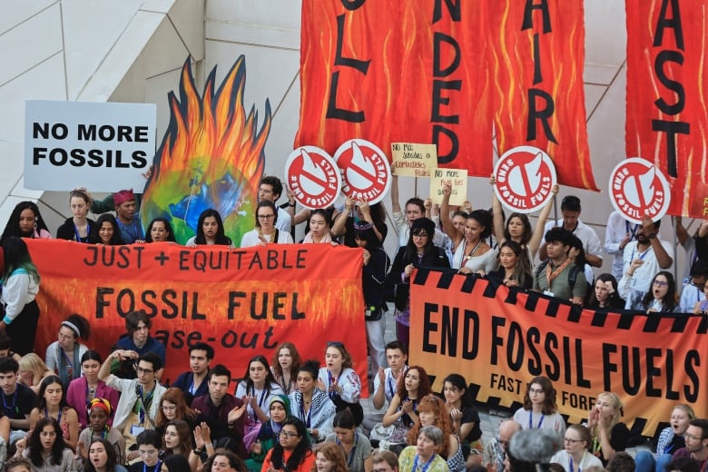 A group of people sitting, holding signs that say 'end fossil fuels' and 'no more fossils'.