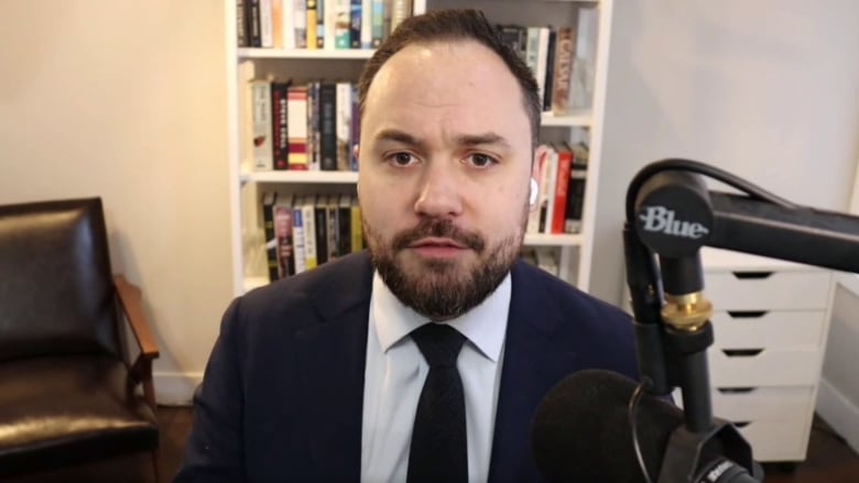 A man wearing a suit sits in front of a bookshelf.