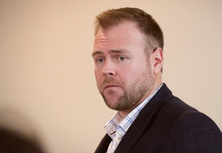 A man in a dark suit and a white-and-checked shirt with a scruffy beard and moustache looks on.