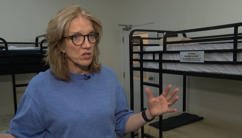 A white blonde woman with glasses and a blue sweater stands in a room with bunkbeds.