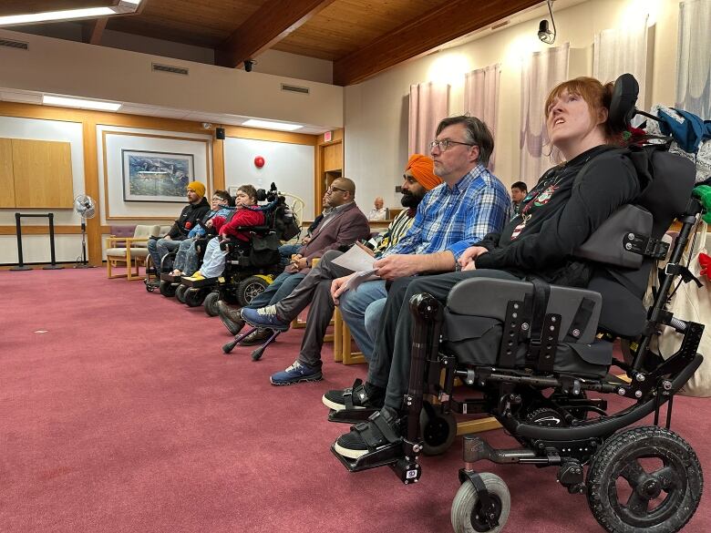 A row of people, some in wheelchairs, sit inside a meeting room.