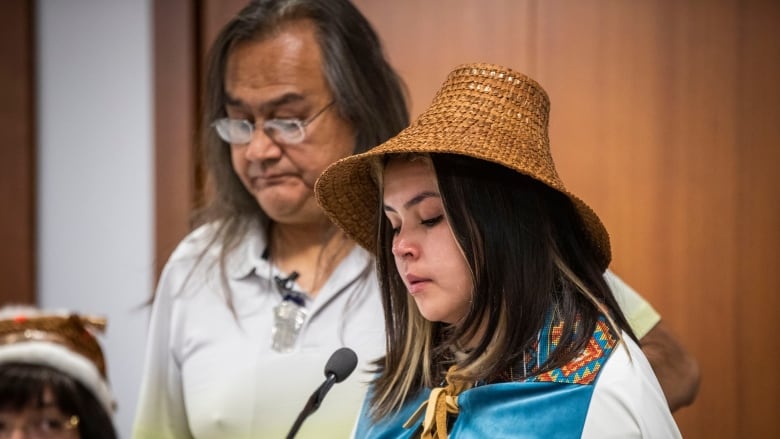 An Indigenous man and a girl wearing Indigenous regalia speak into a microphone while looking down.