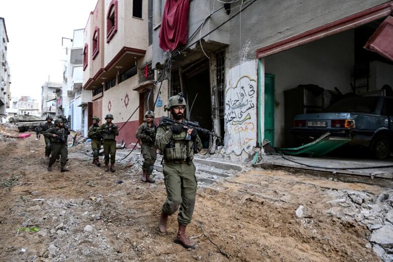 Six Israeli soldiers in green uniforms carry guns as they walk through a ruined street in Gaza.