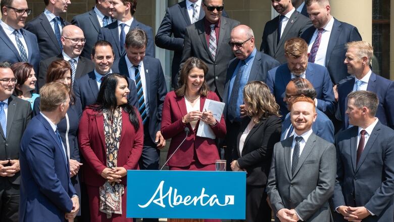 A woman in a burgundy power suit descends stairs to a lectern and fellow politicians gaze warmly at her.