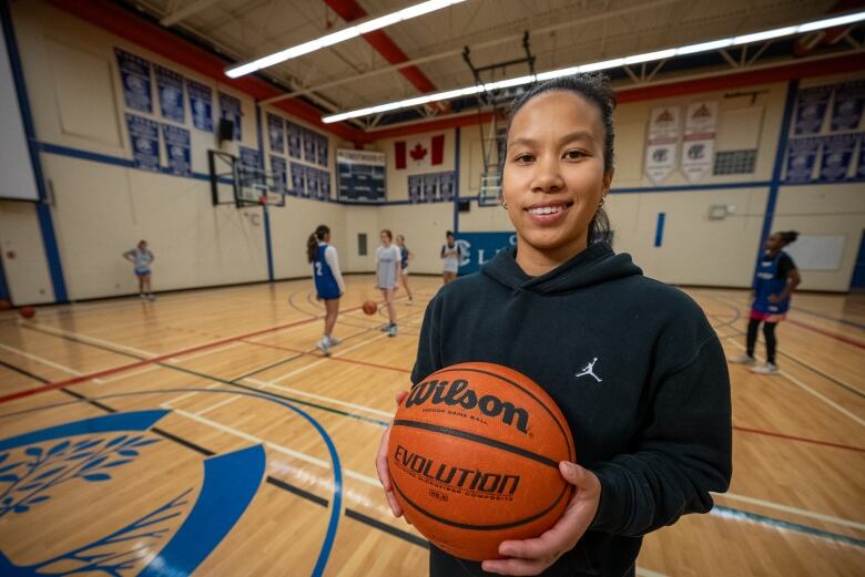 Aprille Deus is pictured holding a basketball while girls practice in the background.