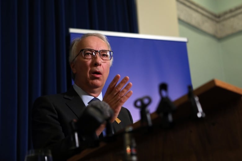 A middle-aged white man stands at a podium wearing a black suit and black-framed glasses.