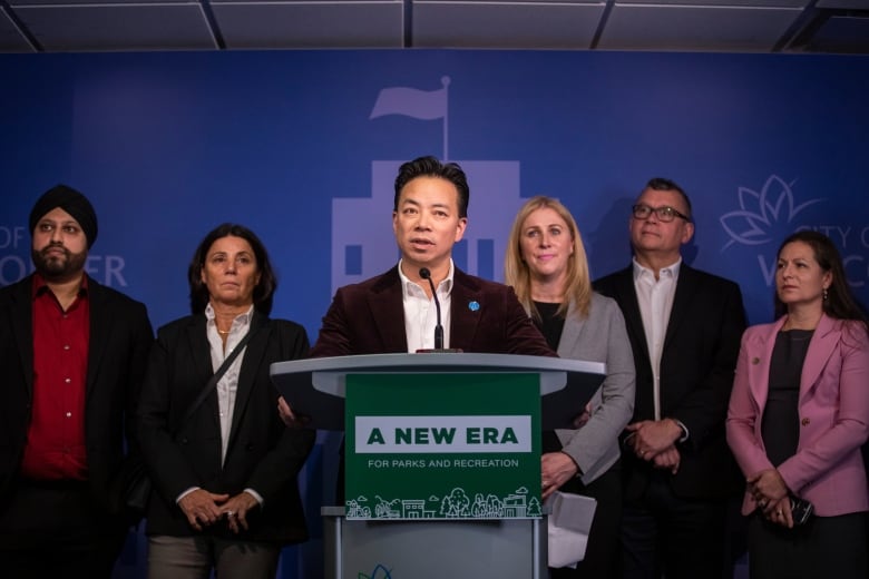 An East Asian man speaks at a podium marked A New Era for Parks and Recreation while being flanked by five other people.