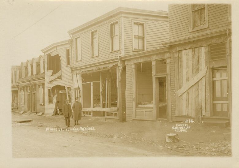 Two men pose in front of buildings that are standing but badly damaged in a black and white photo from 1917.