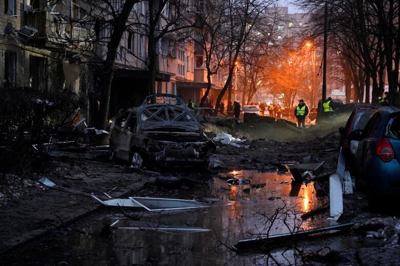 Two people in neon green vests stand amid a destroyed streetscape. A streetlight glows in the distance.