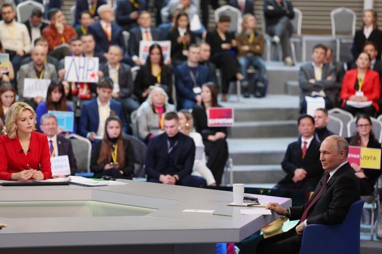 A person sits opposite another person at a large table with an audience in the background.