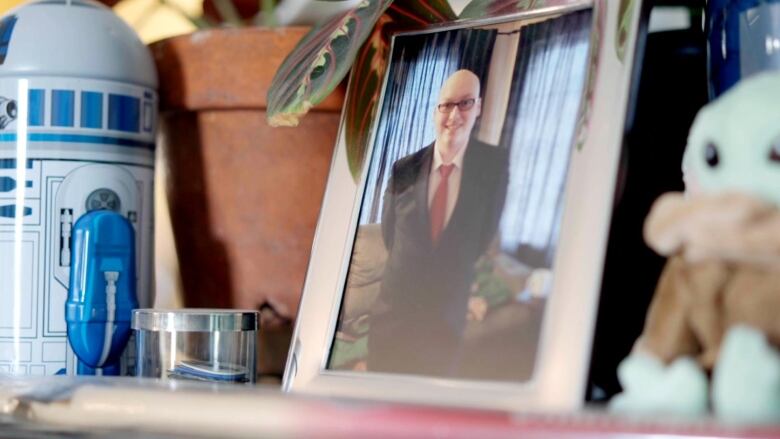 A silver-framed photo of a bald, young man in a dark suit and red tie sits on a desk.  On the right, sits a small stuffed Baby Yoda doll. A clay plant pot sits on the left of the frame with one red and green leaf popping over the top of the pot. To the left of the pot sits a model of R2D2 from Star Wars. 