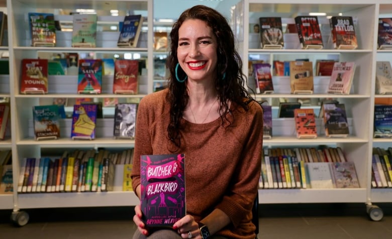 A woman with dark hair holds up a book, Butcher & Blackbird, with library books on shelves behind her.