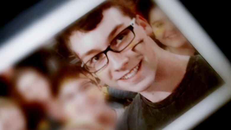 Head and shoulders photo of a young man with short, dark hair smiling. He has dark-framed glasses and is wearing a dark, crew-neck sweat shirt. 