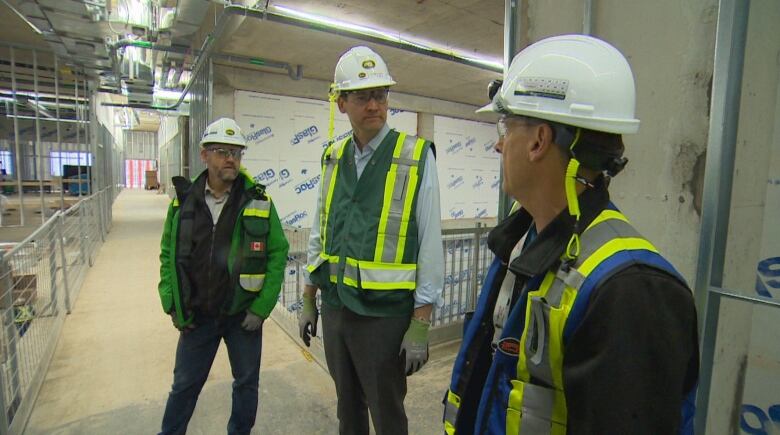 Three men in white construction helmets and high visibility vests stand in a construction site.