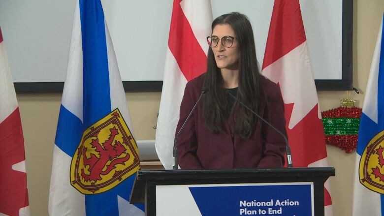 A woman with glasses stands at a podium in front of the Mi'kmaq, Nova Scotian and Canadian flags.