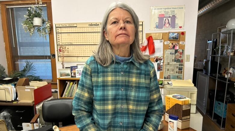 A woman wearing a plaid shirt leans against a desk in an office room.