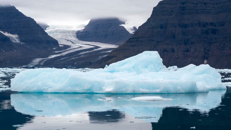 An iceberg is seen floating with mountains and a glacier in the background.