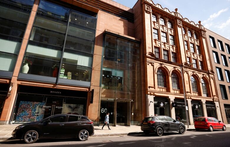 A low-rise building is shown with cars parked and pedestrians walking in front of it.