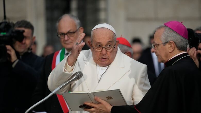 The pope is shown wearing glasses and papal hat speaking into a microphone as a man in religious garments holds an open book in front of him.