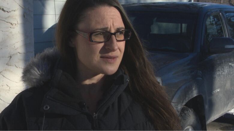 A woman wearing a winter coat stands outside in front of a vehicle in a driveway.