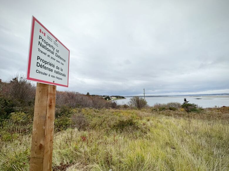 A white sign with black and red lettering saying Property of National Defence sticks up on the shore high above a harbour.