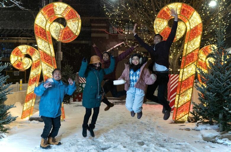 Three people jump in the snow in front of large, lit candy canes.