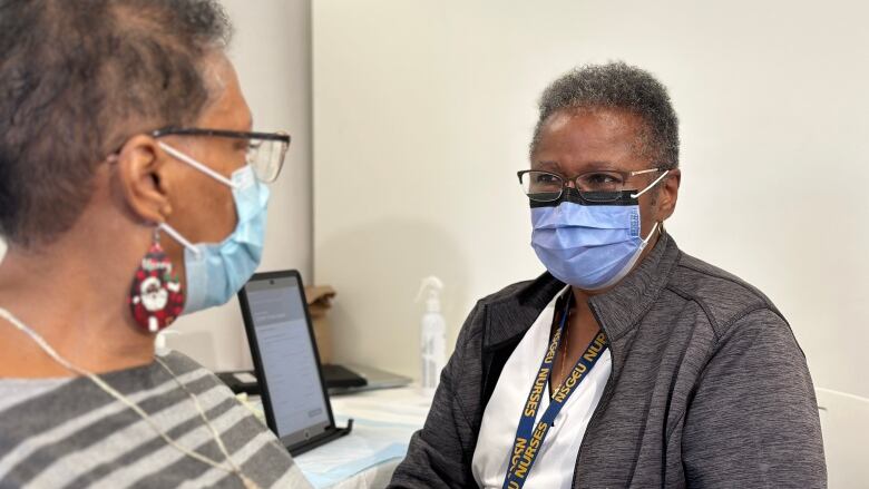 A nurse wearing a surgical mask speaks to a patient, also masked, sitting at a clinic station for administering vaccines.