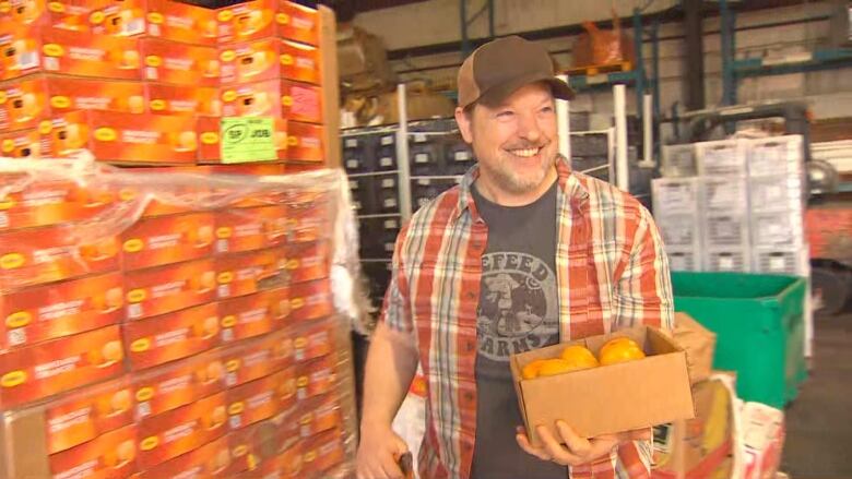 A man stands holding a box of mandarin oranges inside a warehouse full of them.