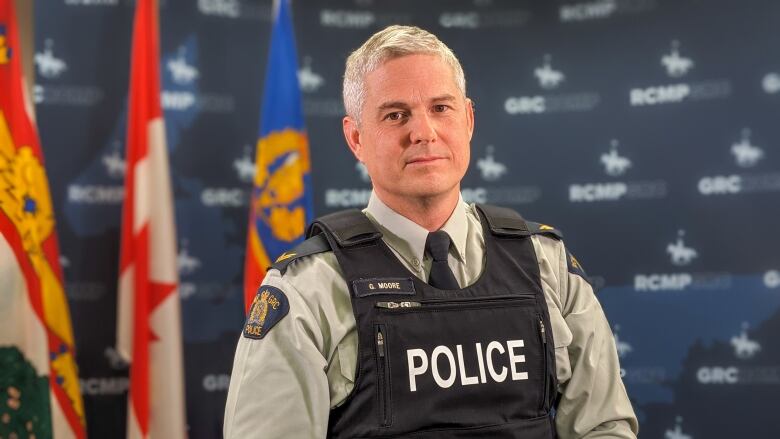 A male police officer in a flack jacket poses in front of flags and an RCMP backdrop.