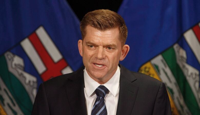 A man wearing a suit and tie stands in front of an Alberta flag.