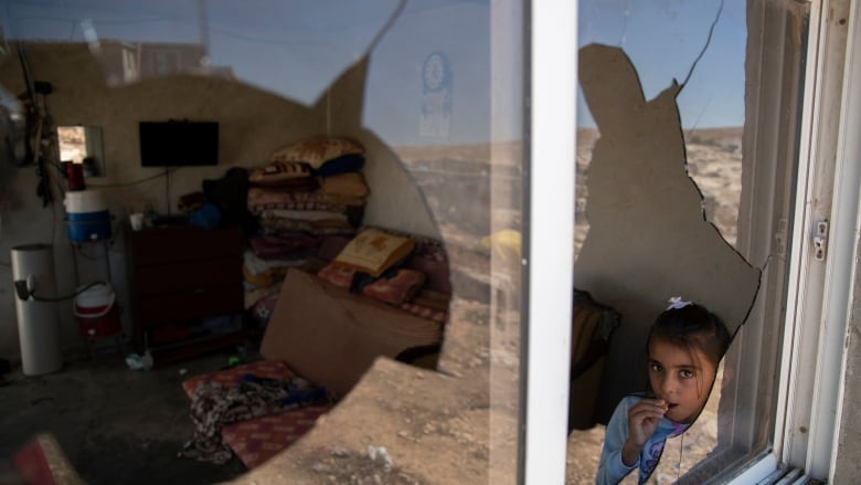 A small girl looks out of a broken window.