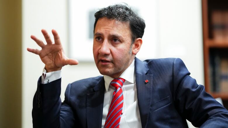 A man in a black tie and red suit gestures with his hand as he speaks.