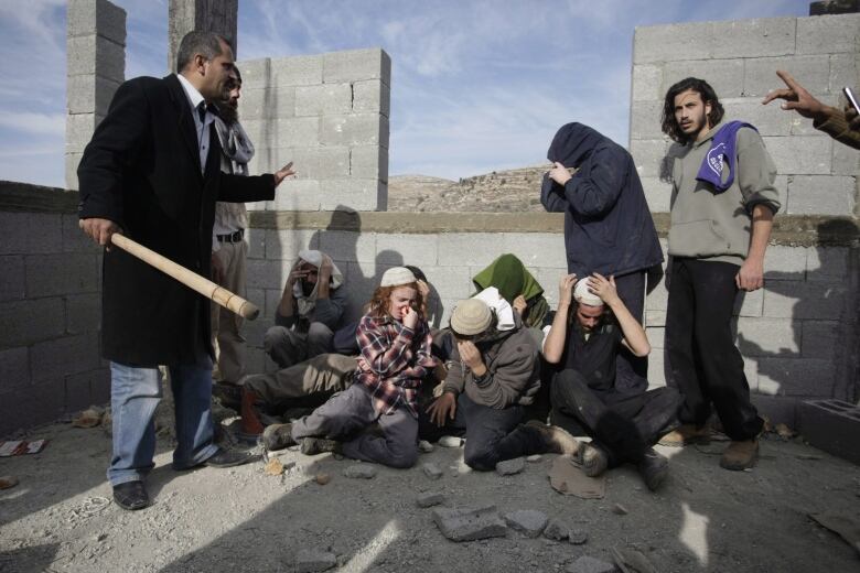 Injured people squat on the ground in an unfinished building, under guard.