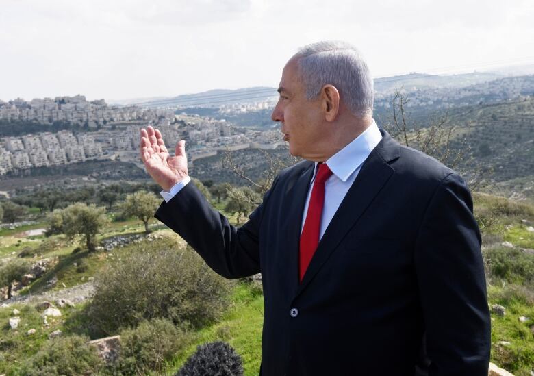 A man in a dark suit looks over a farming valley and gestures.