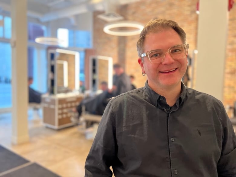 A man with silver hair and thick rimmed glasses is shown in a hair salon.